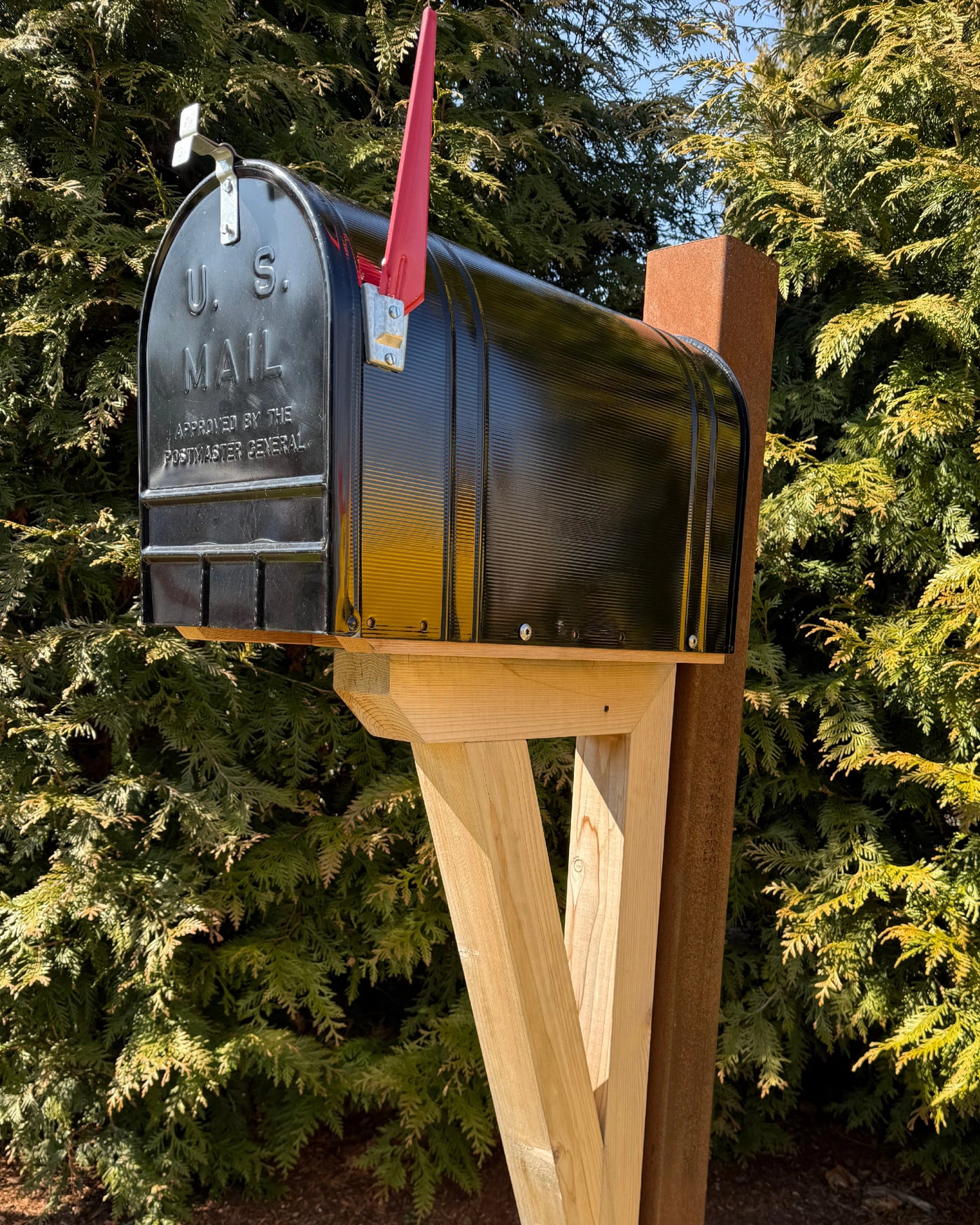Vintage black mailbox mounted on a handcrafted cedar bracket with a weathered steel post outdoors in natural sunlight.