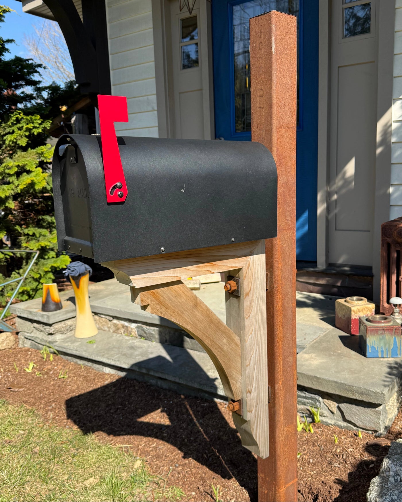 Matte black Titan steel mailbox mounted on a rusted steel post with a handcrafted cedar bracket and bright red flag.