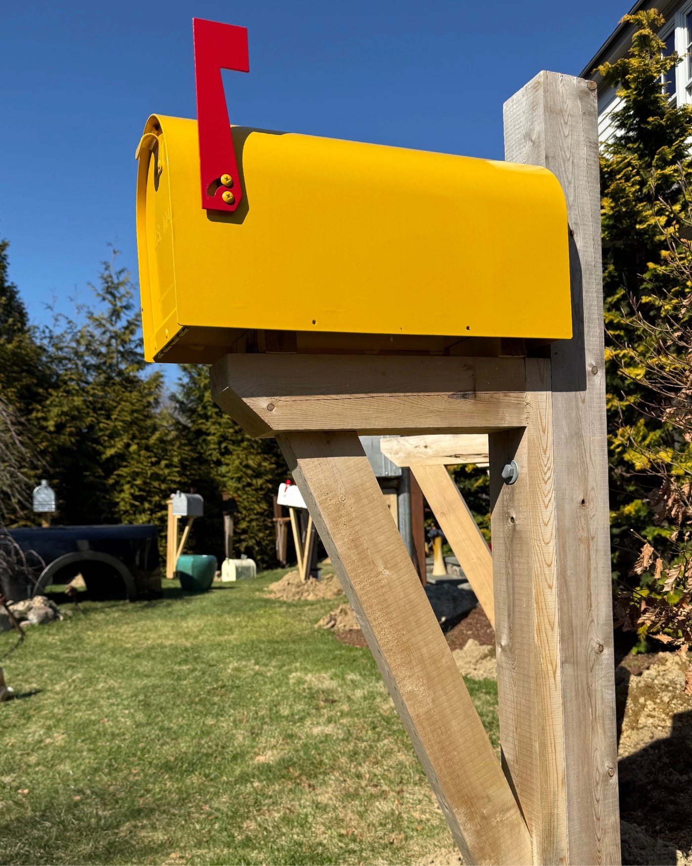 Angled view of a bright yellow Titan mailbox mounted on a white cedar post with handcrafted support bracket detailing.