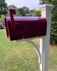Close side view of a burgundy mailbox with matching flag, mounted on a white pine post with a handcrafted curved cedar bracket.