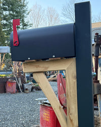 Black vandal-resistant mailbox mounted on a cedar bracket with a steel core post and red mailbox flag outdoors.