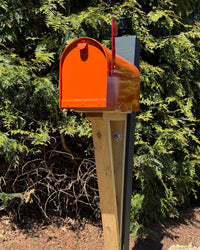 Angled view of a handcrafted orange mailbox set featuring a cedar architectural bracket and durable steel core post surrounded by evergreen trees.