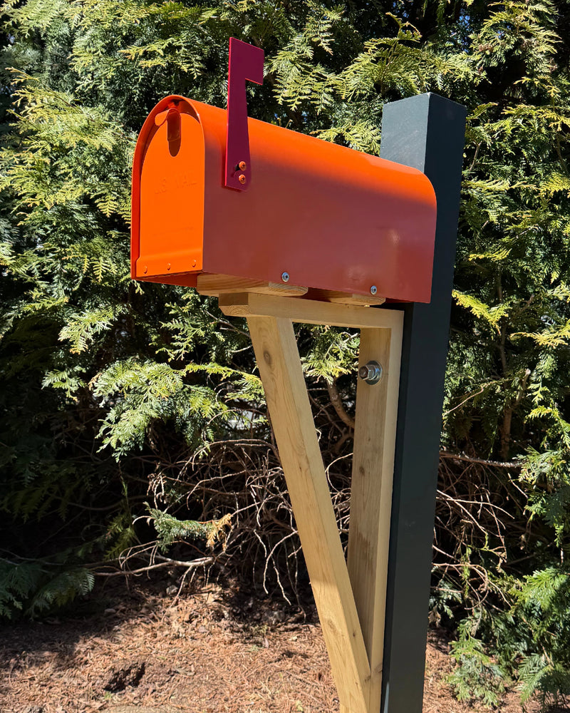 Side view of an orange steel mailbox with natural cedar detailing and a reinforced black steel post in an outdoor setting.
