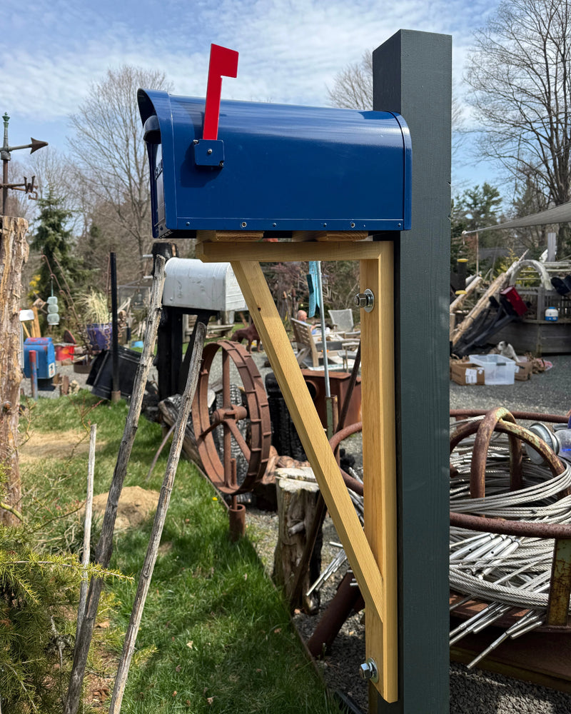 Angled view of a blue modern mailbox with natural cedar detailing and a reinforced steel post in an outdoor setting.