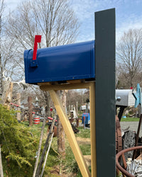 Blue aluminum mailbox mounted on a handcrafted cedar bracket with a black steel core post outdoors in natural sunlight.