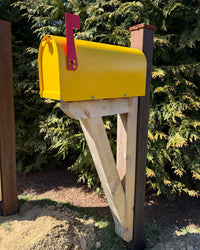 Bright yellow Titan steel mailbox mounted on a handcrafted cedar bracket with a weathered steel post outdoors in sunlight.