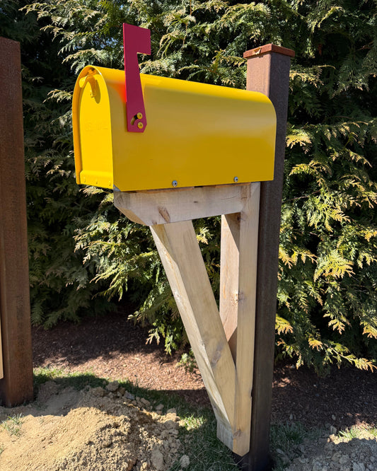 Bright yellow Titan steel mailbox mounted on a handcrafted cedar bracket with a weathered steel post outdoors in sunlight.