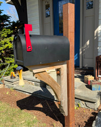 Matte black Titan steel mailbox mounted on a rusted steel post with a handcrafted cedar bracket and bright red flag.