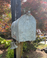 Front angled view of a weathered vintage galvanized mailbox featuring embossed U.S. Mail lettering mounted on a charred cedar post.