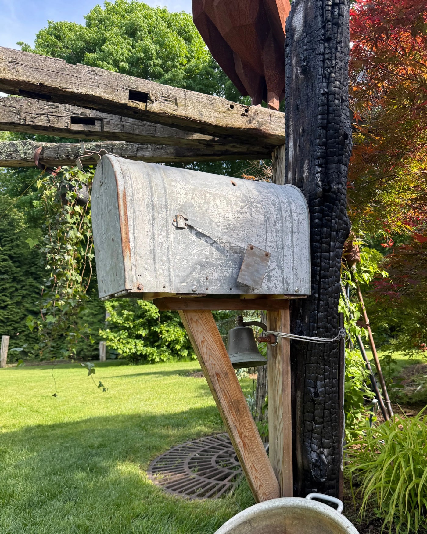 Side angled view of a weathered vintage galvanized mailbox mounted on a charred cedar post with handcrafted wood bracket and rustic bell detail.