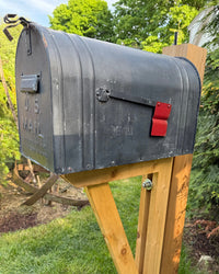 Front angled view of a rustic black U.S. Mail mailbox with red flag and handcrafted cedar support structure.